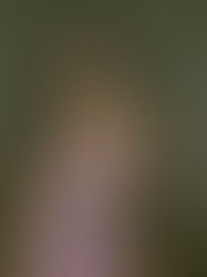 Woman in a lilac dress stands indoors, looking pensive. Dimly lit room with curtain backdrop, lamp and cabinet present.