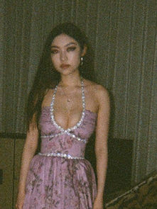 Woman in a lilac dress stands indoors, looking pensive. Dimly lit room with curtain backdrop, lamp and cabinet present.