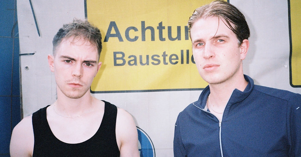 BAUGRUPPE90 stand in front of a yellow sign reading "Achtung Baustelle." They appear serious. One wears a black tank top; the other, a blue jacket.