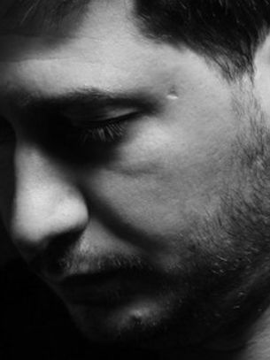 Black and white portrait of a man looking down, wearing a chain necklace. The mood is contemplative, with a dark background.