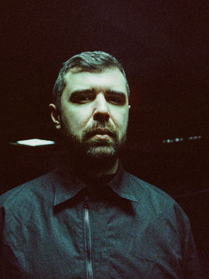 A man in a dark jacket stands in a dimly lit room with red and white lights in the background, creating a moody atmosphere.
