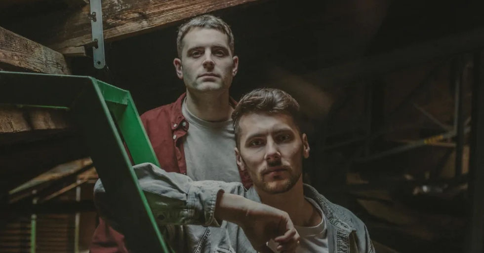 Two serious men stand in a dim warehouse, one in a red shirt behind another in a denim jacket beside green metal framing.