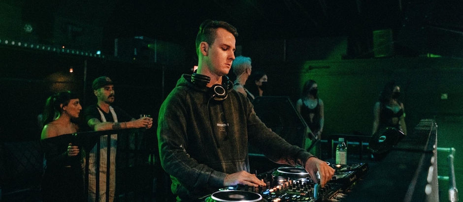 DJ in a dark green-lit club adjusts a mixer while onlookers watch from behind the booth.