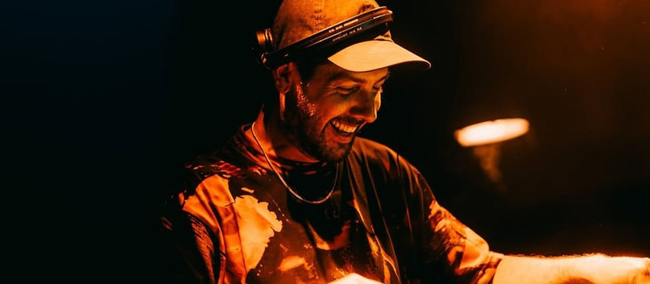 A DJ in a cap and headphones smiles while performing. Warm lighting casts an orange glow, creating a lively, energetic atmosphere.
