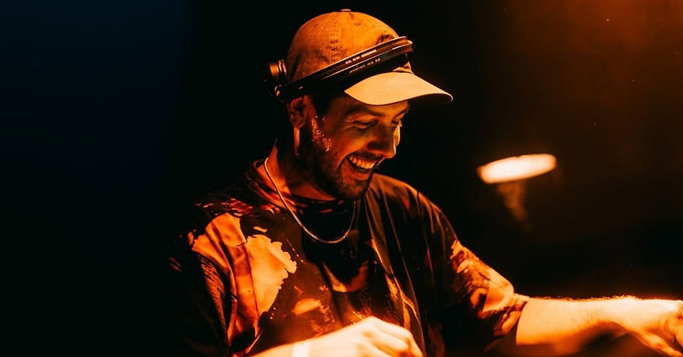 A DJ in a cap and headphones smiles while performing. Warm lighting casts an orange glow, creating a lively, energetic atmosphere.