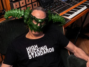 Man reclines in a studio chair wearing a green monster mask and a black shirt reading HIGH FUCKING STANDARDS, near keyboards.