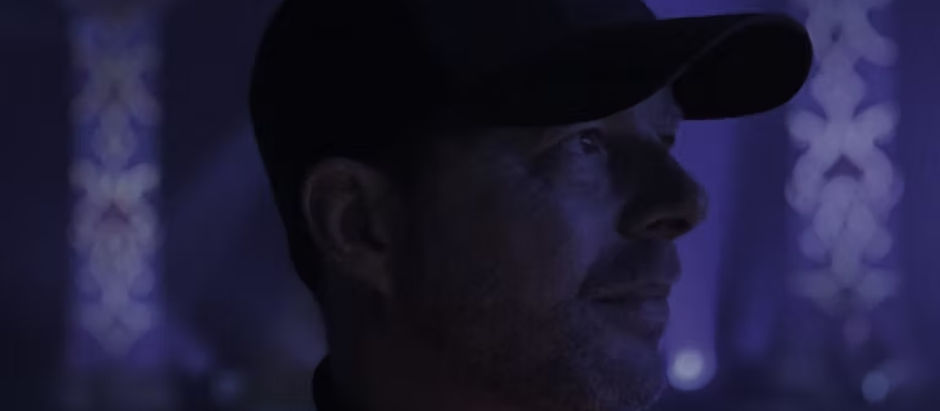 Man in a cap looking contemplative, bathed in purple light with blurred decorative columns in the background, creating a serene mood.