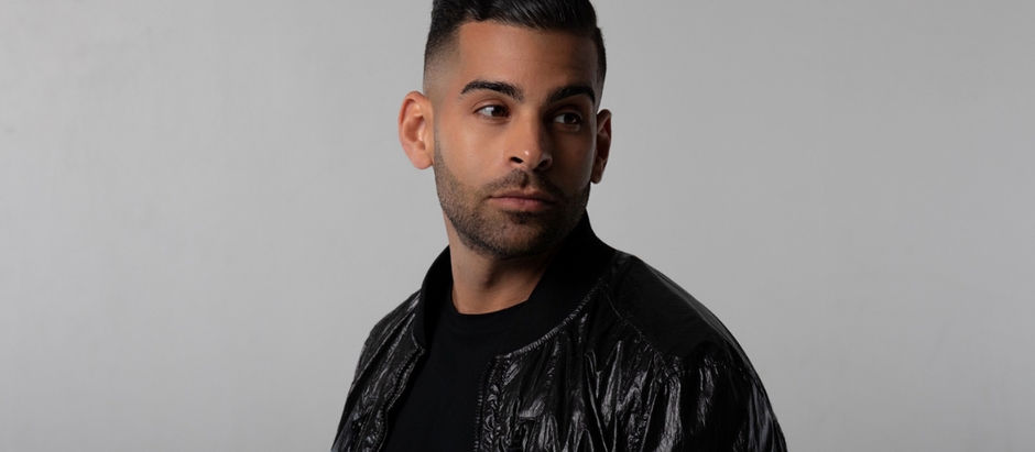Man in a black shiny jacket looks to the side against a plain gray background, conveying a contemplative mood.
