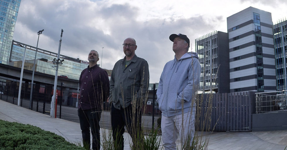 Three men stand outdoors in a modern city plaza under cloudy skies, looking upward in a calm, contemplative mood.