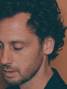 A man with curly hair looks down pensively against a warm, orange background. He wears a dark shirt, and the mood is introspective.
