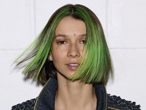 Portrait of a woman with neon green bob hair and a denim jacket against a plain white background, looking calm.