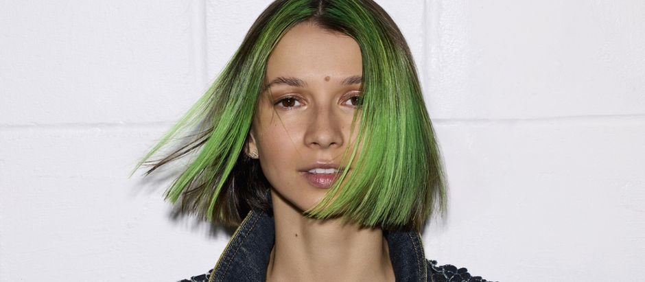 Portrait of a woman with neon green bob hair and a denim jacket against a plain white background, looking calm.