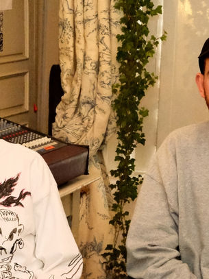 Two men seated in a room with music equipment, one with long hair and graphic shirt, the other in a cap. Calm atmosphere, patterned curtain.