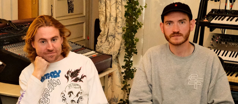 Two men seated in a room with music equipment, one with long hair and graphic shirt, the other in a cap. Calm atmosphere, patterned curtain.