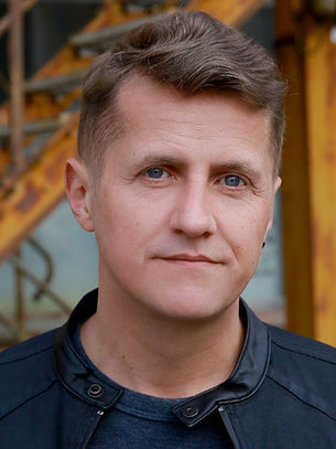Man in black jacket stands in front of rusty orange staircase, with a brick wall in the background. He appears calm and composed.