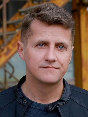 Man in black jacket stands in front of rusty orange staircase, with a brick wall in the background. He appears calm and composed.