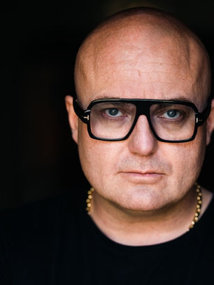 Bald man with black glasses and a gold necklace stares intently at the camera, set against a dark background, creating a serious mood.