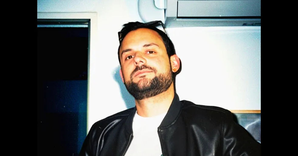 Bearded man in a black jacket poses indoors against a white wall and dark doorway.