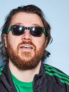 Bearded man with sunglasses, wearing a green-striped jacket, stands against a light blue background, appearing relaxed.