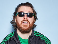 Bearded man with sunglasses, wearing a green-striped jacket, stands against a light blue background, appearing relaxed.