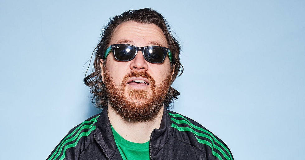 Bearded man with sunglasses, wearing a green-striped jacket, stands against a light blue background, appearing relaxed.