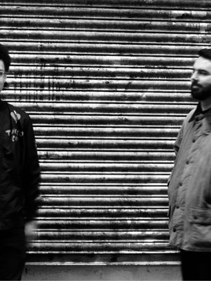 Two men stand before a corrugated metal shutter in black and white, one facing camera, the other in profile, moody street scene.