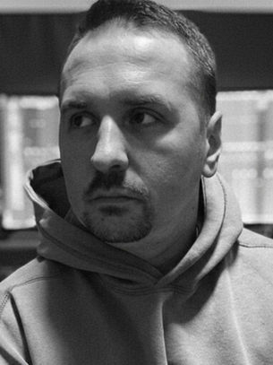 Man in hoodie in a sound studio with monitors and speakers in the background. Black and white, serious expression.