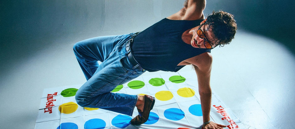 Man in black tank top and jeans playing Twister, balancing on a colorful mat with green, yellow, and blue dots. Energetic mood.