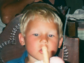 Blond young boy in a restaurant chair eating a frozen treat, with diners and tableware blurred behind him.