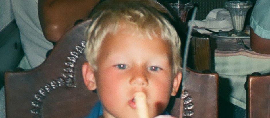 Blond boy in a restaurant sits at a table, eating a popsicle or ice cream with a focused, calm expression.