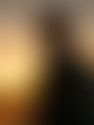 Bearded man in cap and glasses stands on a rooftop at sunset, backlit by a glowing orange sky, with a contemplative mood.
