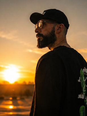 Bearded man in cap and glasses stands on a rooftop at sunset, backlit by a glowing orange sky, with a contemplative mood.