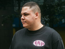 Man in a black shirt with a purple emblem stands outdoors, looking to the side. Background is blurred with greenery and buildings.