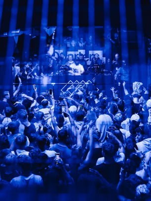 Crowd inside a blue-lit club, people dancing and cheering, DJ at the front with equipment. Energetic and lively atmosphere.