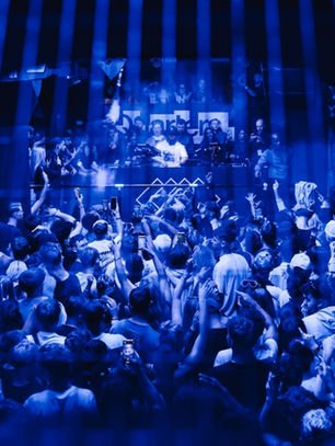 Crowd inside a blue-lit club, people dancing and cheering, DJ at the front with equipment. Energetic and lively atmosphere.