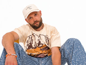 Bearded man in a beige cap and dog graphic T-shirt sits against a white background, wearing speckled blue jeans.