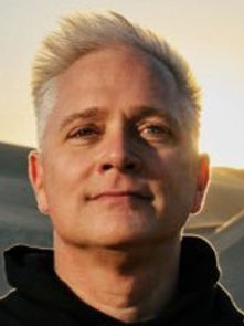 Man with short blond hair stands in a sunlit desert, wearing a black top. The background features sand dunes and a warm, orange sky.