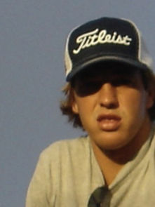Young man outdoors wearing a blue "Titleist" cap and gray shirt, with a neutral expression. Clear blue sky in the background.
