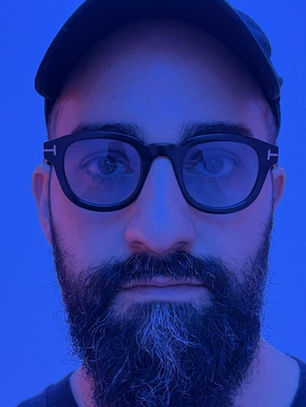 Close-up of a bearded man in glasses and a cap, lit in blue with a neutral expression against a plain blue background.