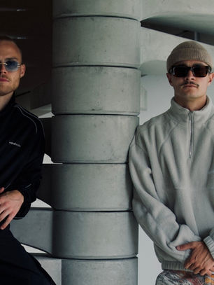 Two men in sunglasses pose indoors by a concrete staircase, one in black and one in gray, with a cool, moody vibe.