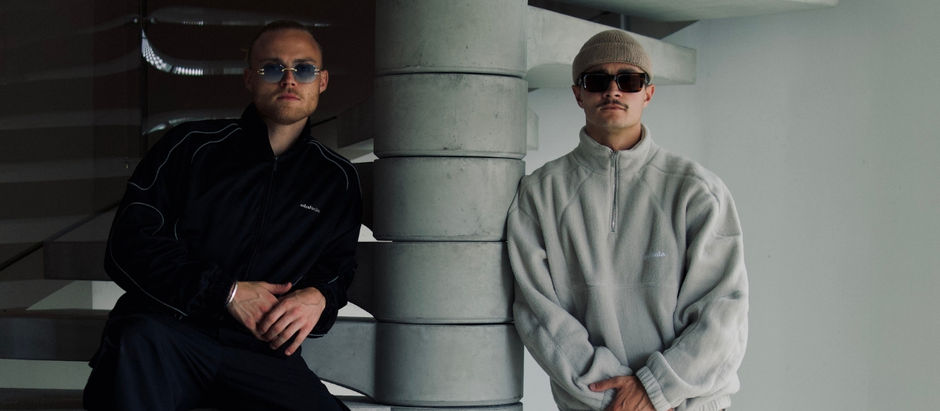 Two men in sunglasses pose indoors by a concrete staircase, one in black and one in gray, with a cool, moody vibe.