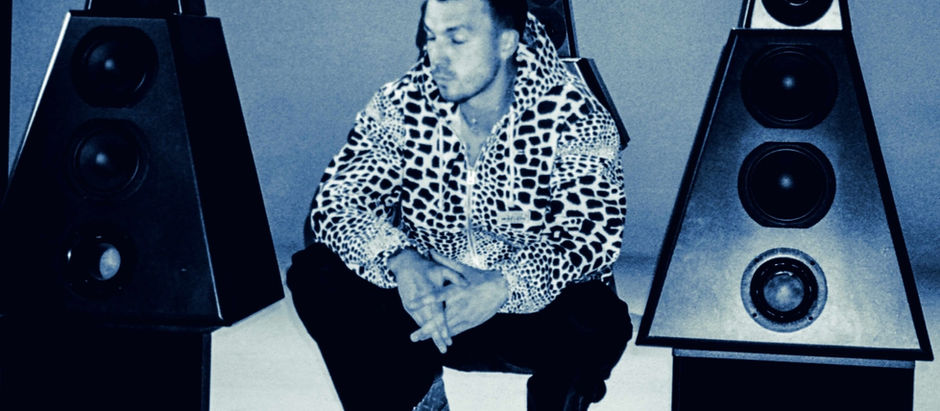 Man in leopard-print hoodie squats between two large speakers, in a moody blue-toned setting, appearing contemplative.