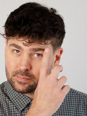 Close-up of a bearded man in a checkered shirt touching his temple against a plain gray background, looking serious.