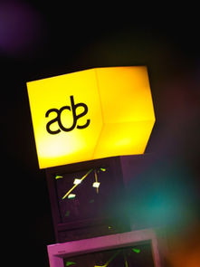 Glowing yellow cube with "ade" text atop stacked monitors. Colorful blurred lights in foreground create dynamic, vibrant atmosphere.