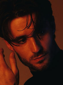 A man in a dark shirt poses against a brown backdrop. His gaze is pensive, with dramatic lighting highlighting his face and tousled hair.