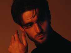 A man in a dark shirt poses against a brown backdrop. His gaze is pensive, with dramatic lighting highlighting his face and tousled hair.