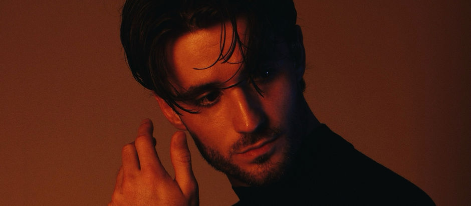 A man in a dark shirt poses against a brown backdrop. His gaze is pensive, with dramatic lighting highlighting his face and tousled hair.