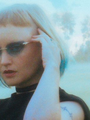 A person with sunglasses and a contemplative look stands in a misty field under a cloudy sky. Blue hair and a tattoo are visible.