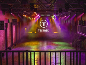 Empty club stage lit in purple, pink, and gold, with TESTBED logo glowing above a reflective floor.