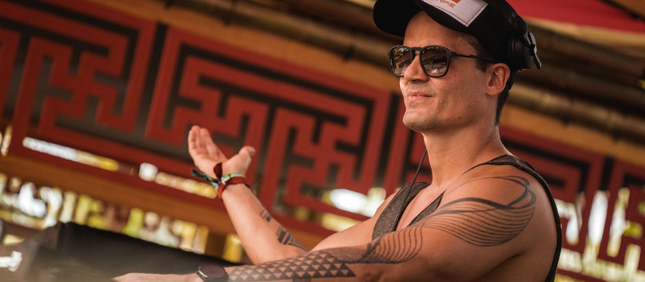 DJ in a cap and sunglasses playing music outdoors. He wears a tank top, showing tattooed arms, against a patterned red and black backdrop.
